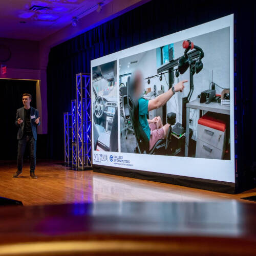 Paul Fink, assistant professor in the College of Computing, gives remarks on stage at Loosemore Auditorum, large screen showing adaptive vehicle behind him